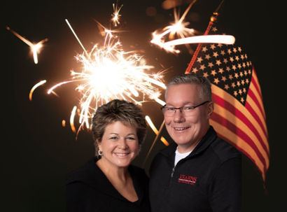 General Manager Diane Kearns and Business President Greg Kearns standing in front of a sparkler and an American flag